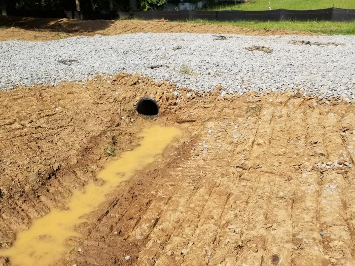 Sandy drainage ditch with muddy water flowing into a black culvert under gravel