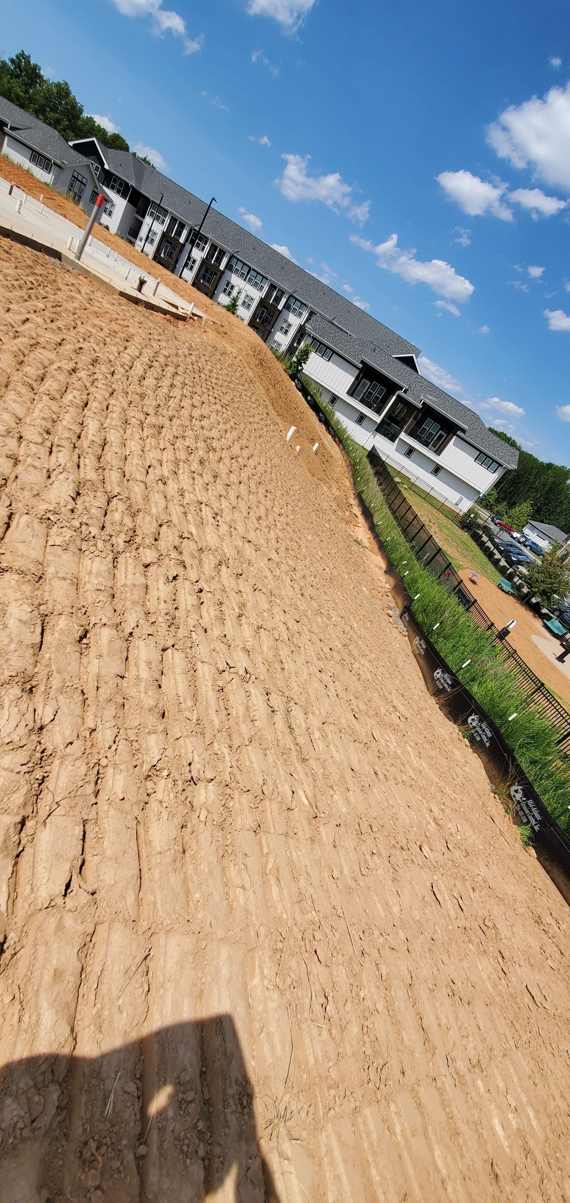 Tilted view of a dirt construction site beside white buildings under a blue sky with clouds