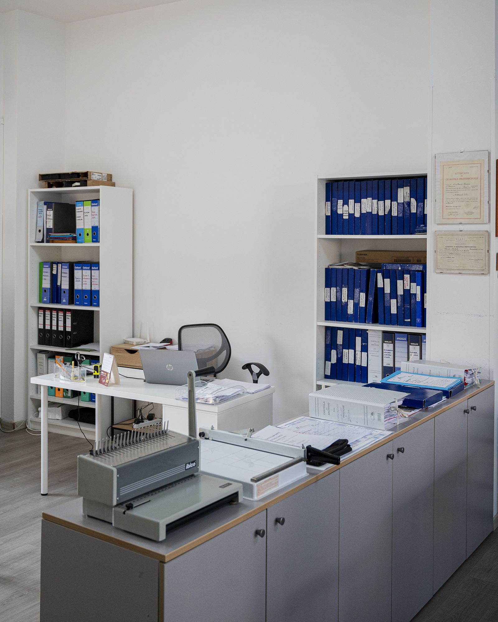 Ufficio con pareti bianche, scrivanie, scaffali pieni di raccoglitori e una rilegatrice.