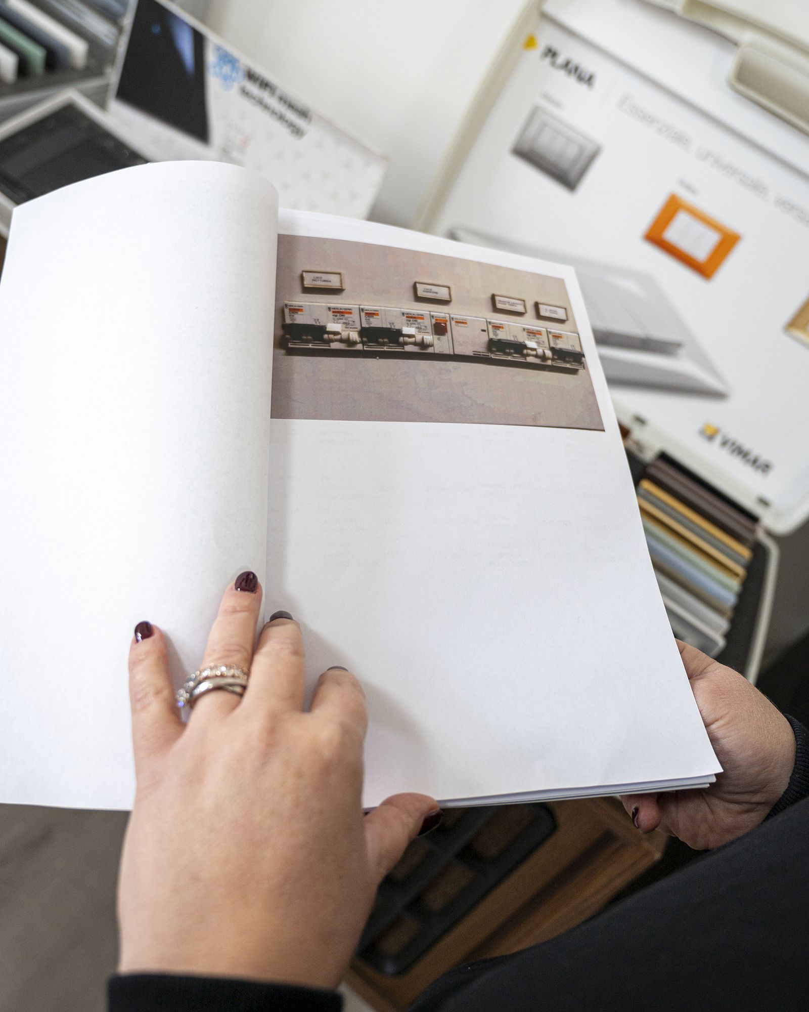 Persona che tiene un libro aperto e mostra una foto di interruttori elettrici. Sullo sfondo, altre brochure di prodotto.