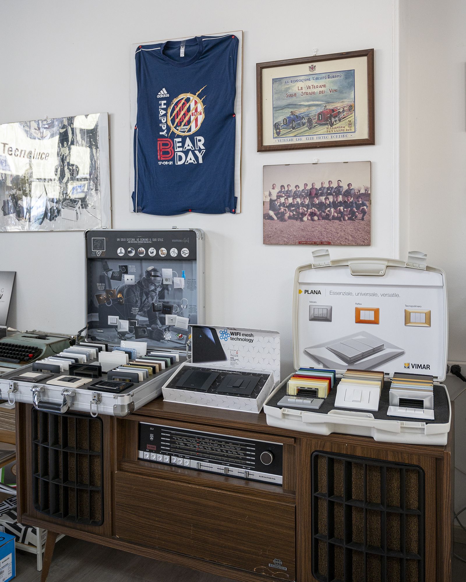 Una consolle in legno espone apparecchiature audio, tra cui un mixer, registratori a cassette e opere d'arte incorniciate appese a una parete bianca.