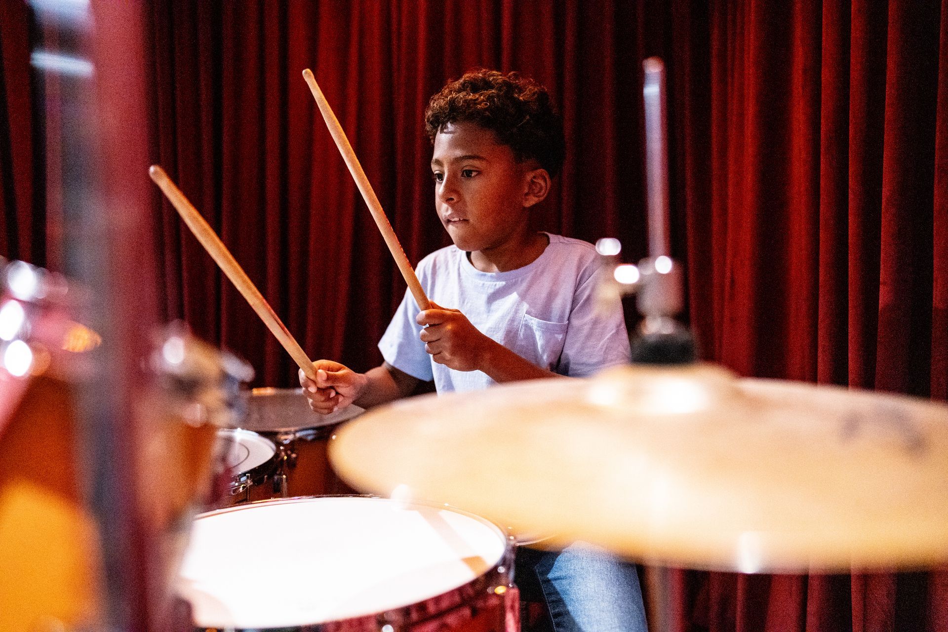 A young boy is playing drums on a stage in front of a red curtain. A young boy is playing drums on a stage in front of a red curtain.