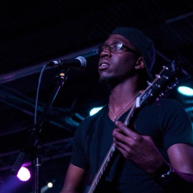 Man playing guitar and singing into a microphone on stage, under blue and purple lights. Man playing guitar and singing into a microphone on stage, under blue and purple lights.