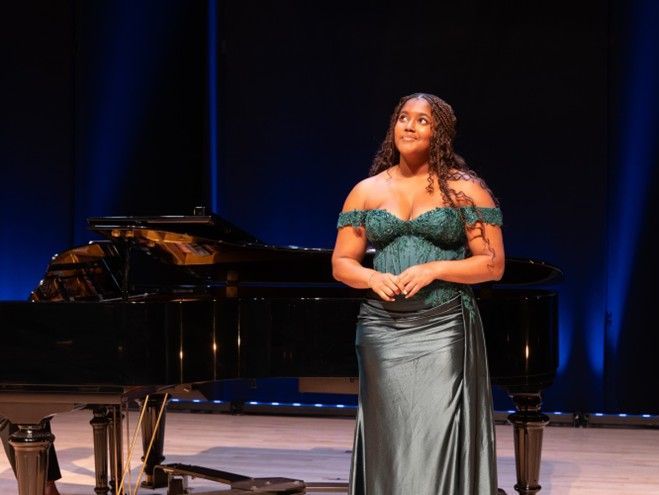 Woman in green dress on stage next to piano, looking upward, smiling. Blue backdrop. Woman in green dress on stage next to piano, looking upward, smiling. Blue backdrop.