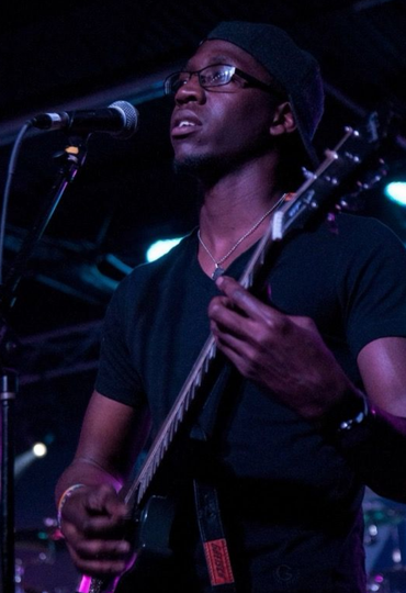 Man playing a guitar and singing into a microphone on stage, wearing a black shirt and a beanie. Man playing a guitar and singing into a microphone on stage, wearing a black shirt and a beanie.