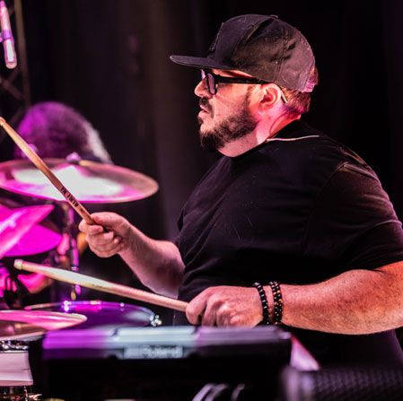 Drummer playing drums on stage, wearing a black cap and sunglasses, with a purple-lit drum kit. Drummer playing drums on stage, wearing a black cap and sunglasses, with a purple-lit drum kit.