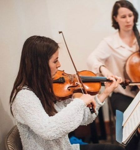 Woman playing violin, focused, with another woman playing cello in the background. Light-colored clothing, indoor setting.
