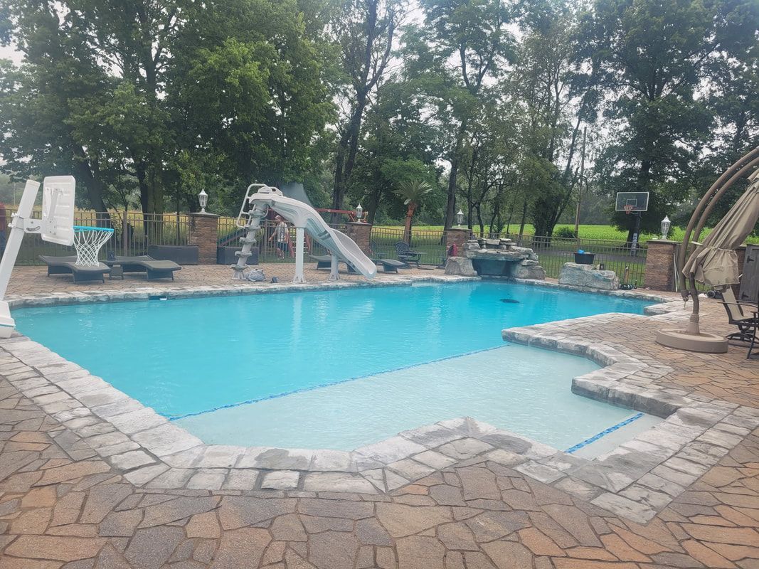 Pool with a slide, basketball hoop, and stone surround in a yard with trees.
