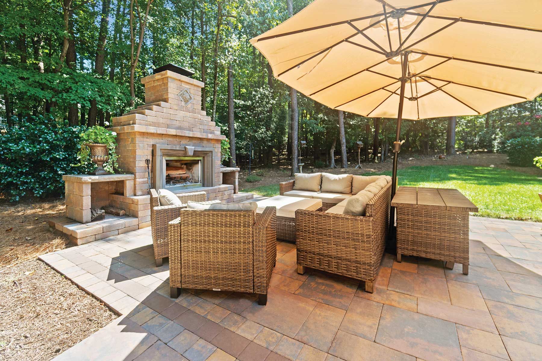 Patio with outdoor fireplace, seating, and umbrella in a wooded backyard setting.