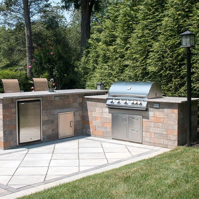 Outdoor kitchen with grill, refrigerator, and storage cabinets on a stone patio.