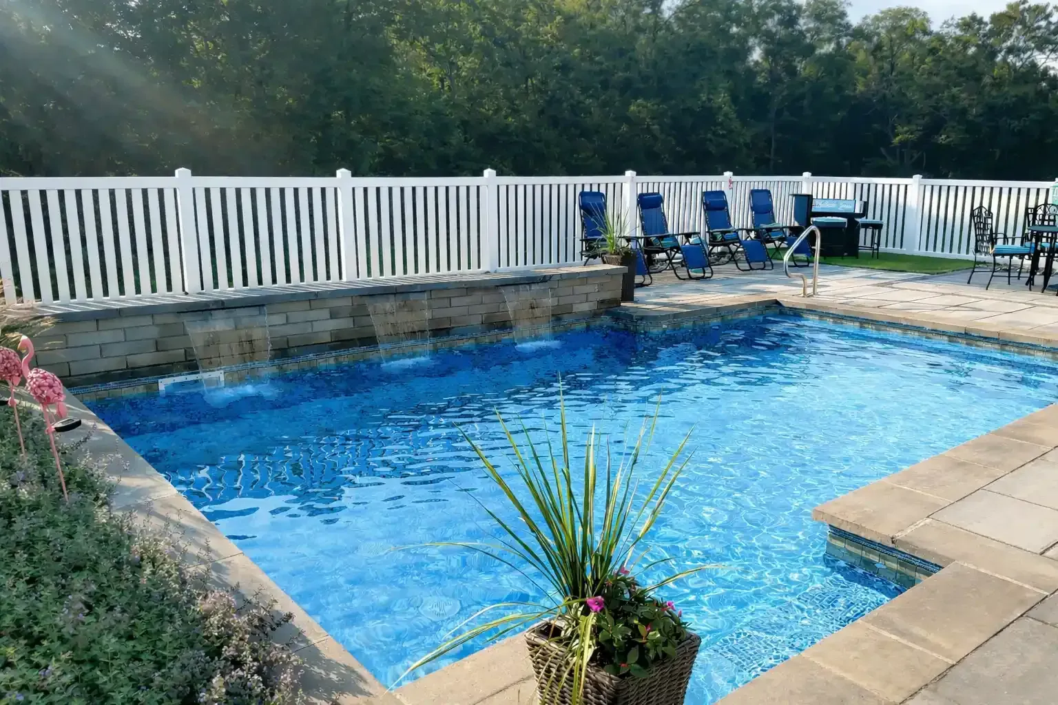 Swimming pool with waterfall feature, surrounded by a stone deck and white fence, chairs in the background.