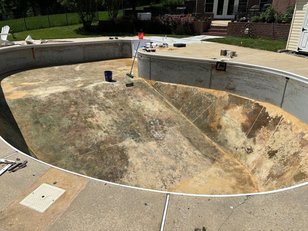 Empty, oval-shaped swimming pool with stained interior and concrete border. Surrounded by patio and house in background.