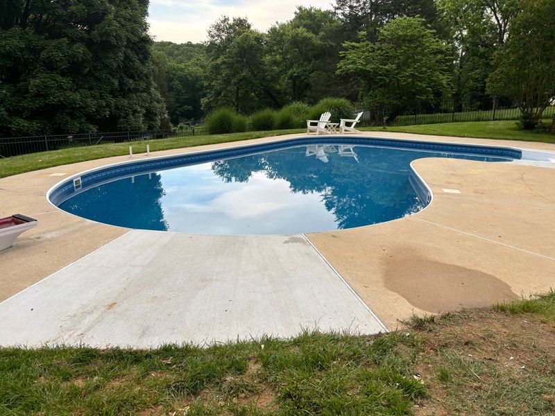 Pool with blue water and concrete deck, white chairs, and trees in background.