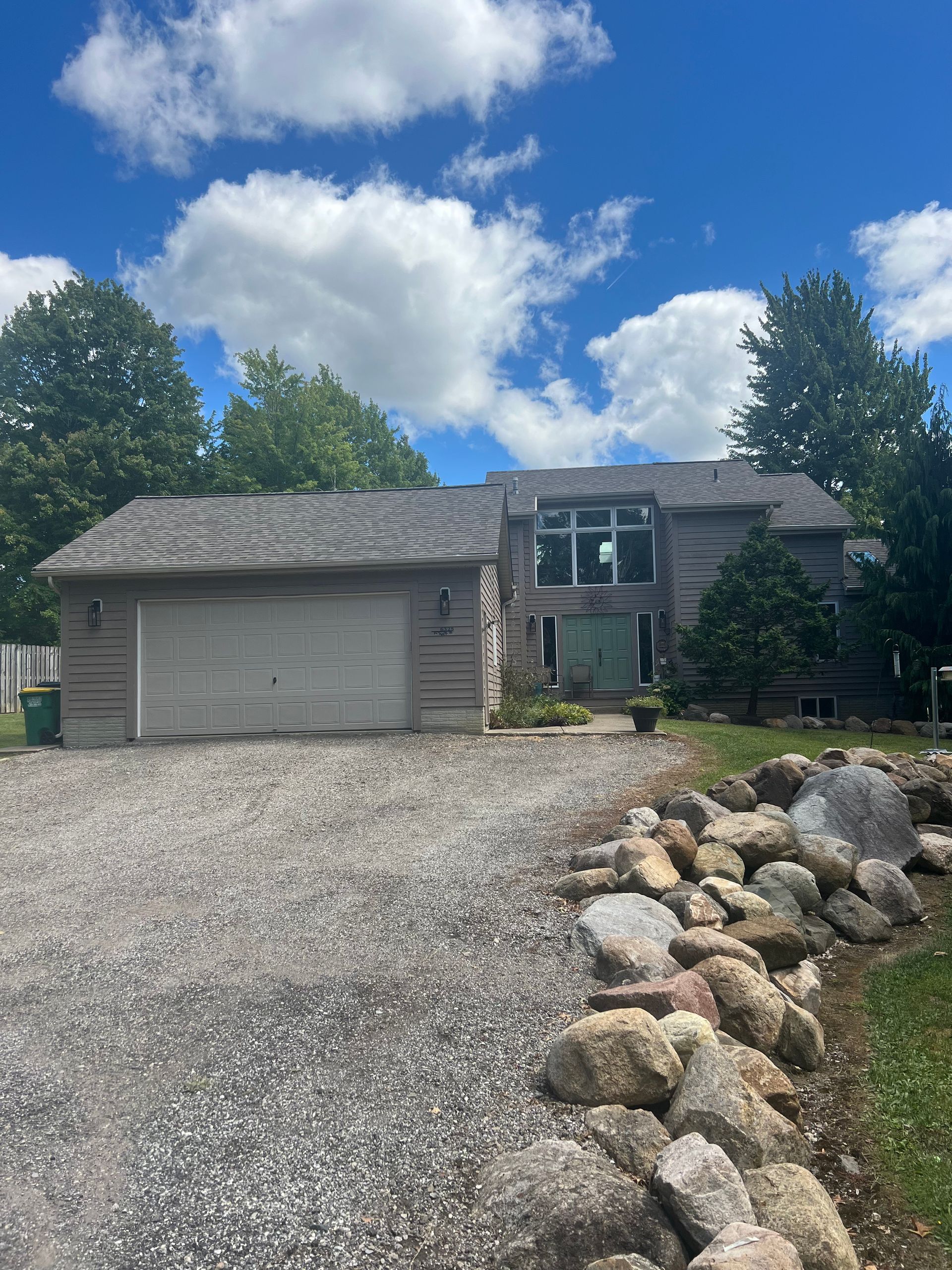 Gray, wood-sided house with attached garage. Gravel driveway. Blue sky with white clouds.