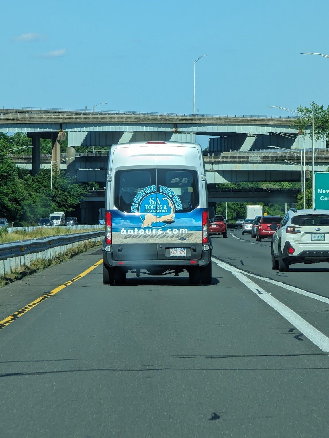 A van with a blue and white wrap drives on a highway, viewed from behind, with an overpass visible in the background.