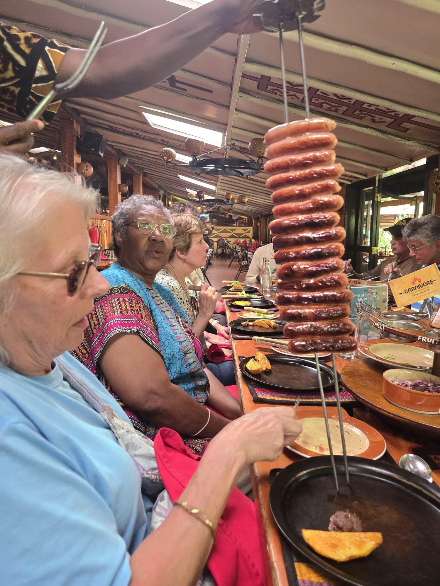 A server holds a tall skewer of cooked sausages over a plate at a dining table where several people are eating.