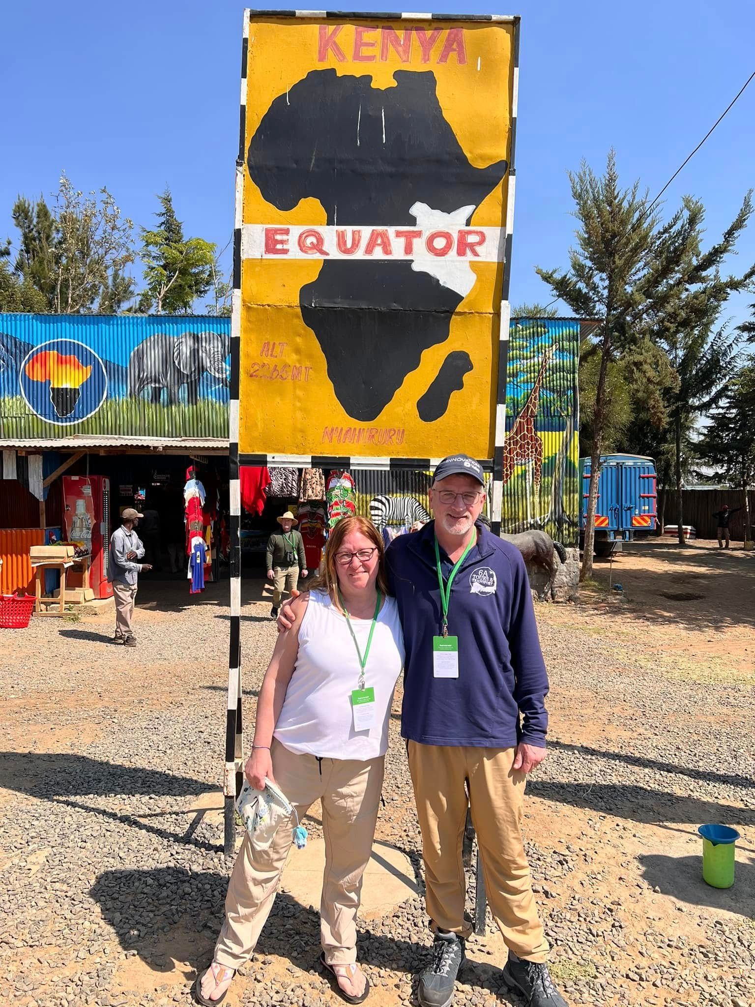 Two people stand in front of a yellow sign marking the Equator in Kenya, with a painted map of Africa in the center.