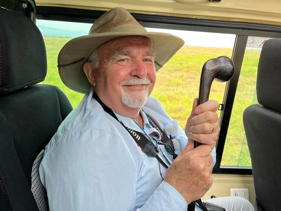 A smiling person in a tan hat and blue shirt sits in a vehicle, holding a dark, knobbed wooden walking stick.