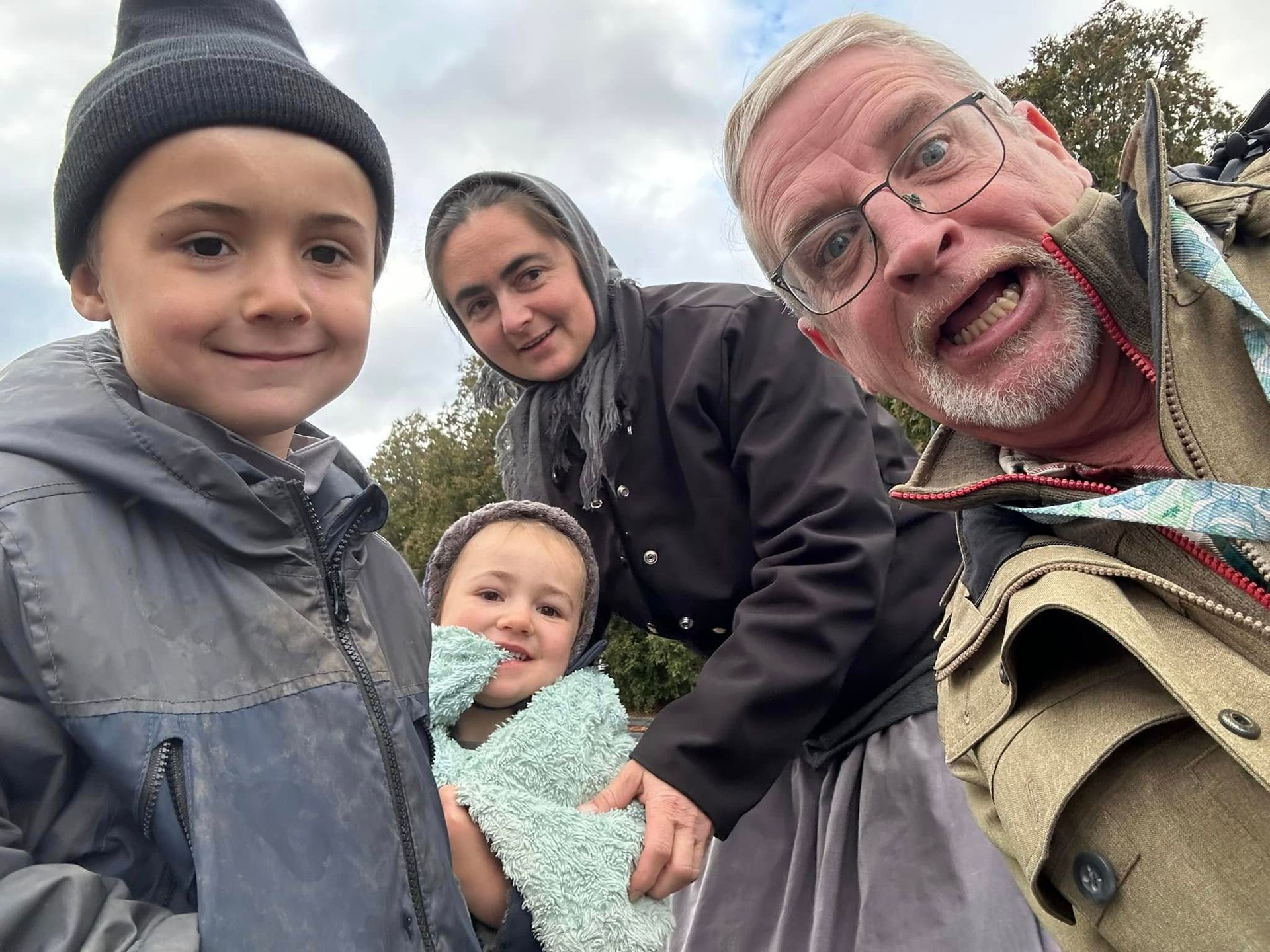 A selfie of four people outdoors, including a man with glasses making a silly face, a woman, and two smiling children.