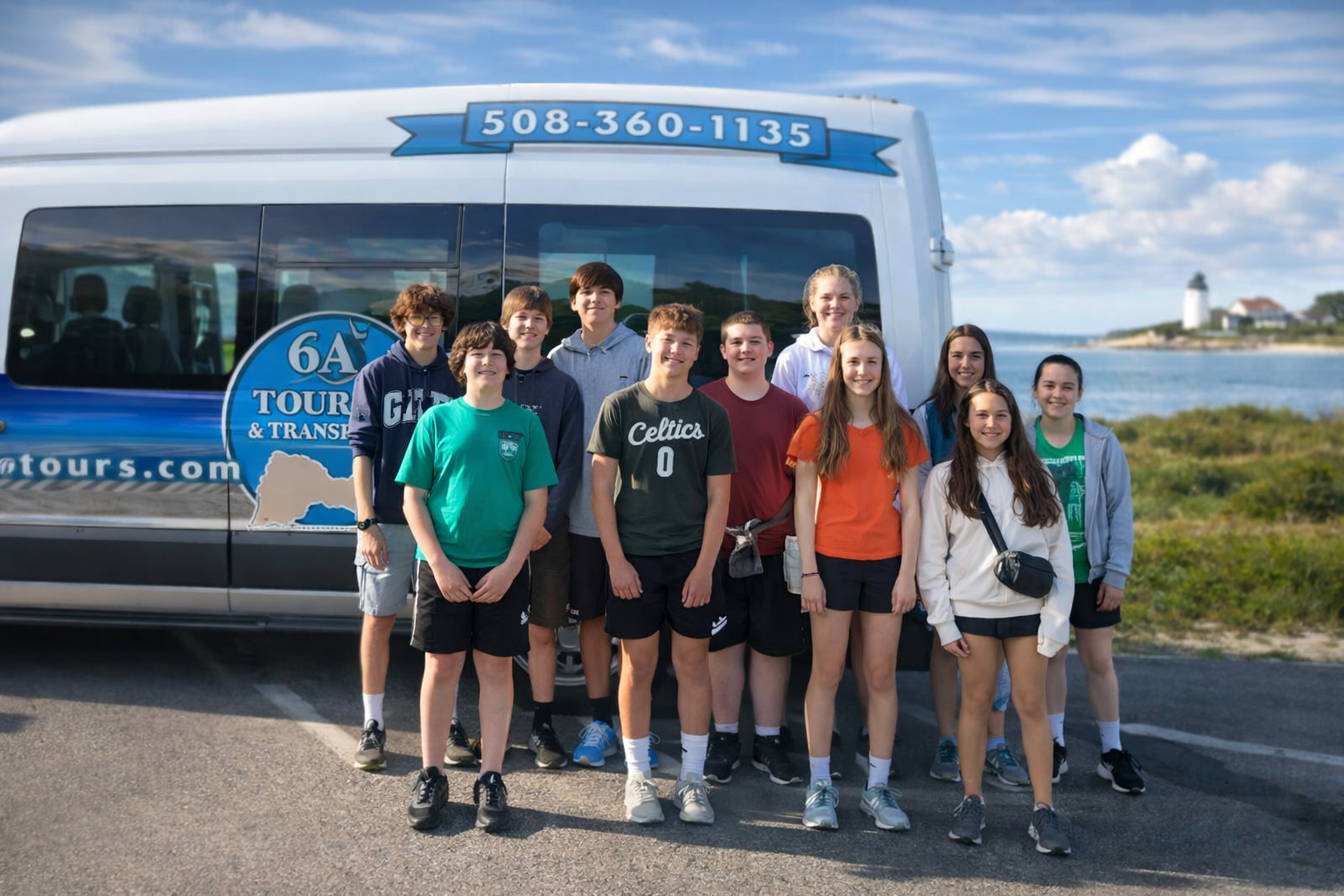 A group of eleven people standing in front of a 6A Tours and Travels van by a coastline and lighthouse.