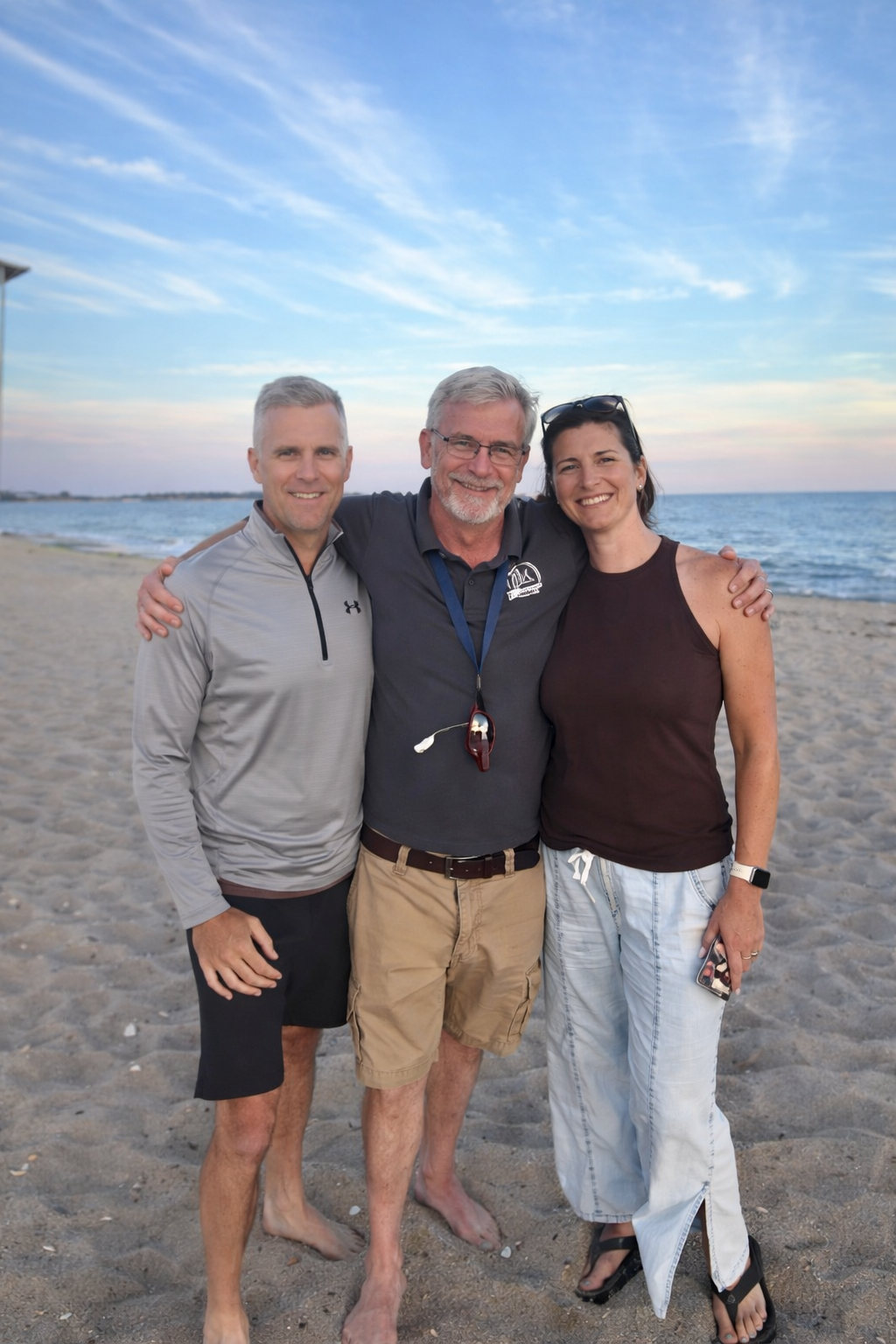 Three people stand together on a sandy beach at sunset, smiling with their arms around each other.