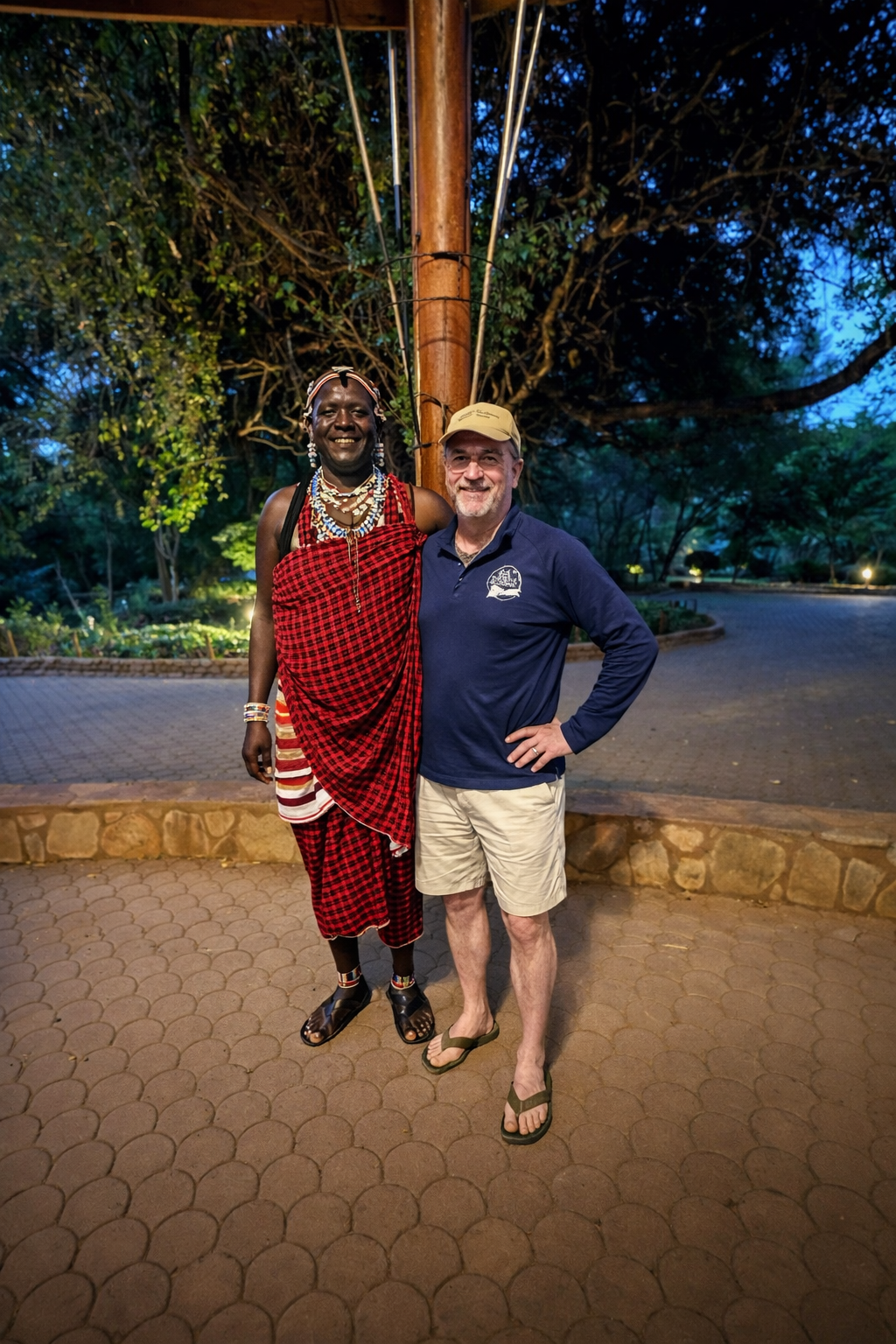 A person in traditional Maasai attire and a person in a polo shirt and shorts stand together outside at twilight.