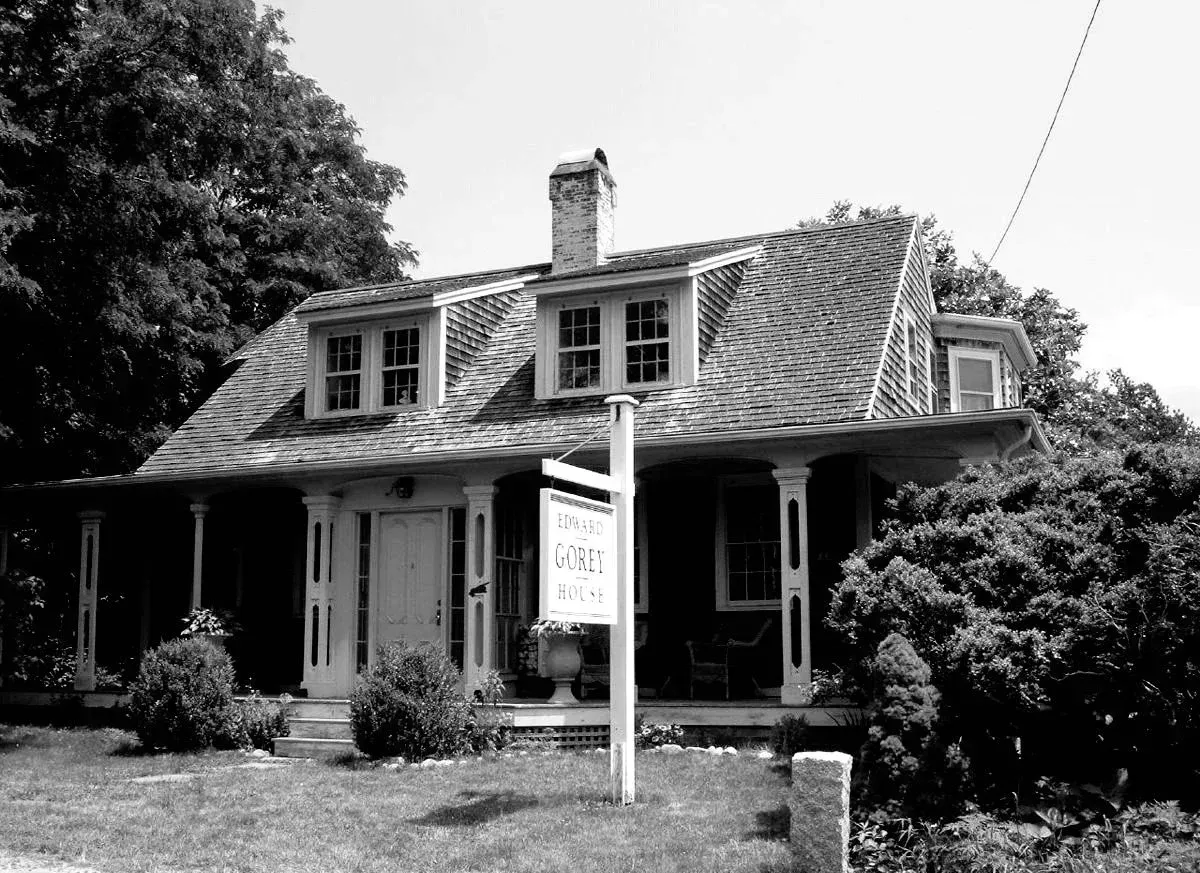 A black-and-white view of a historic cottage-style house with a covered front porch and a real estate sign in the yard.