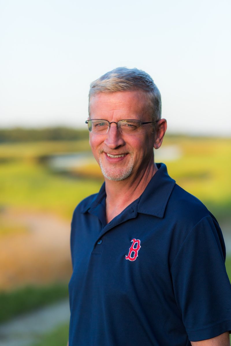 A smiling person with glasses and a navy Boston Red Sox polo shirt stands outdoors in front of a blurry, grassy landscape.