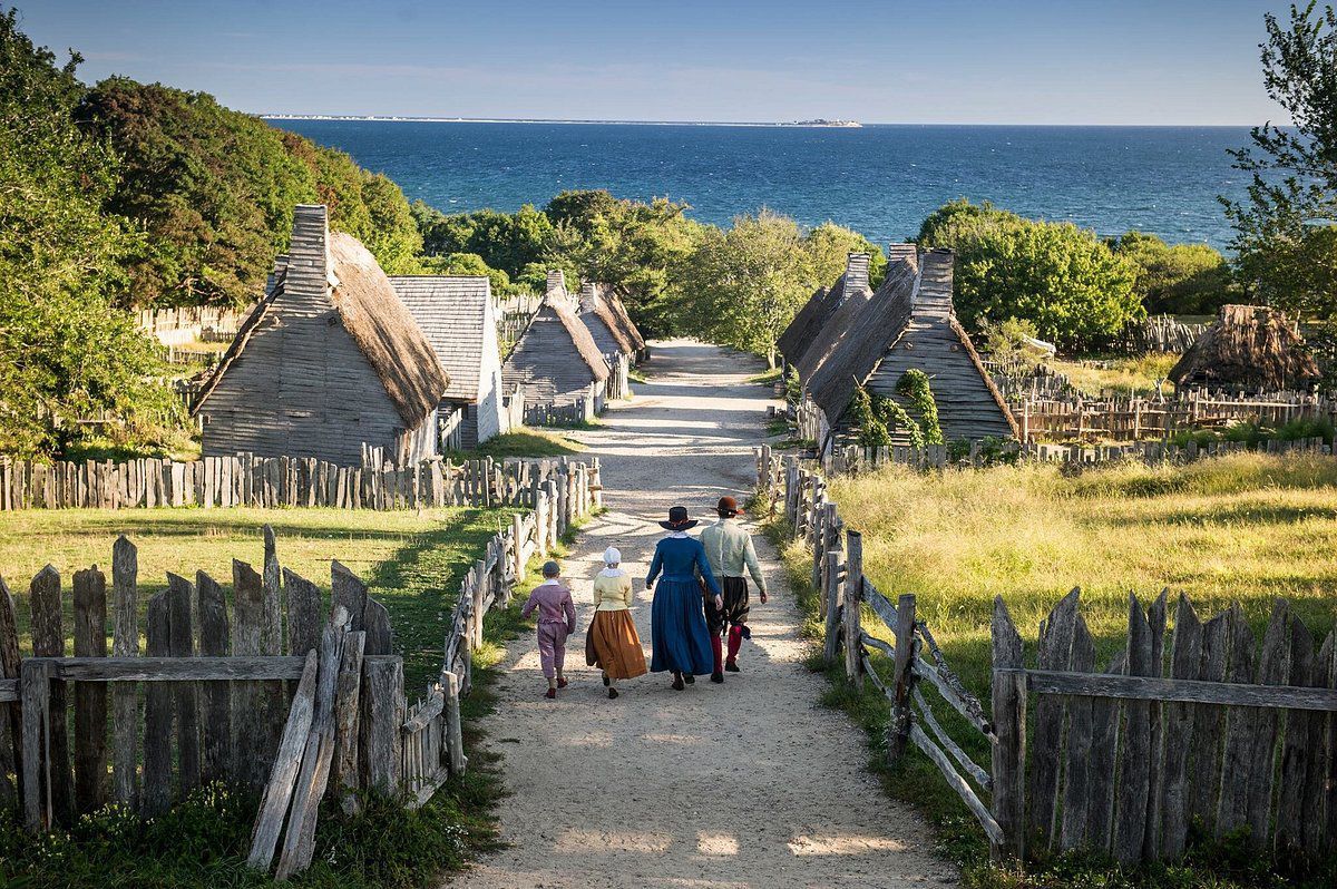People walking down a dirt path between rustic cottages toward the sea on a sunny day.