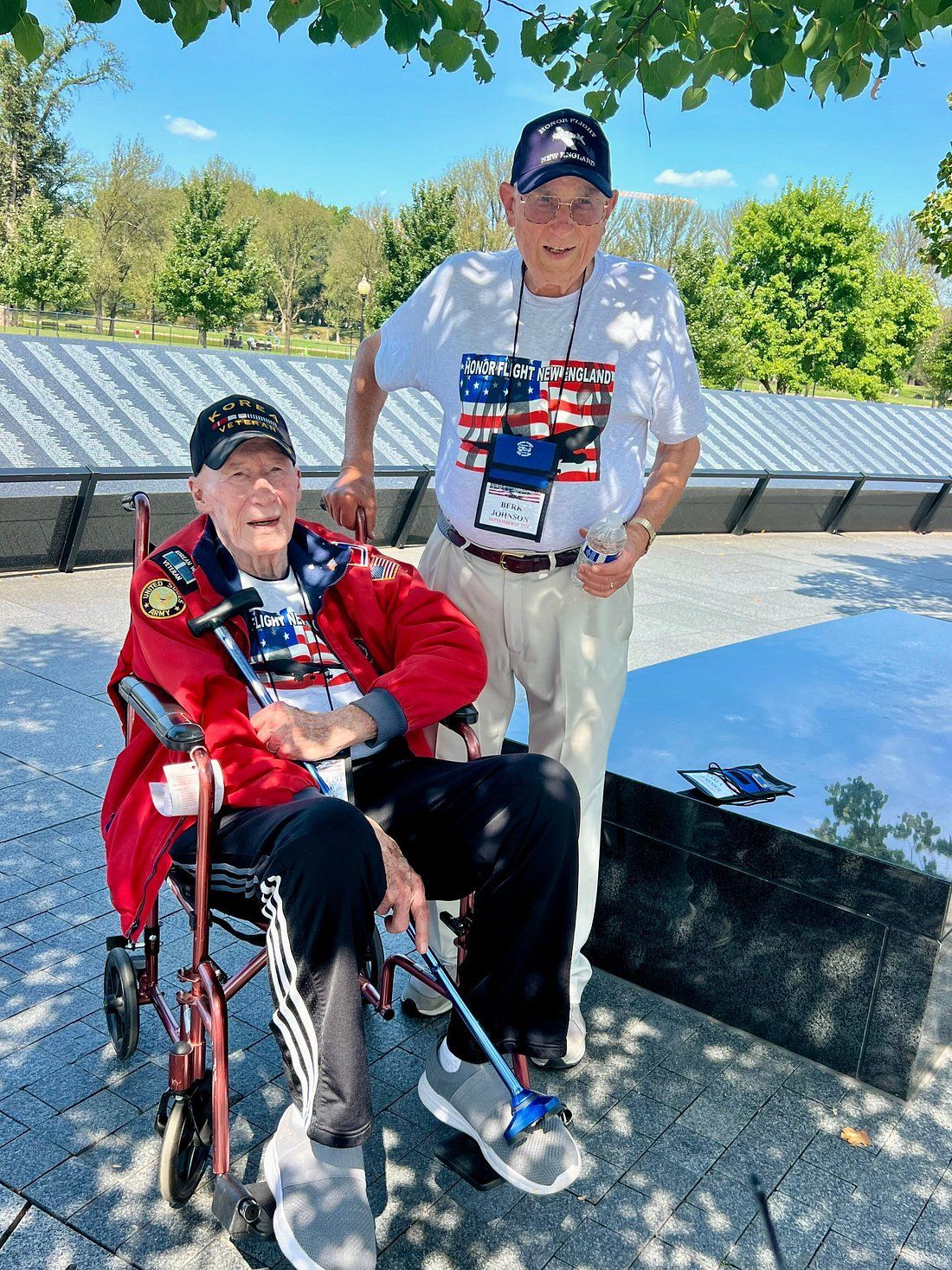 Two veterans standing and sitting in a wheelchair by a stone memorial wall in a sunny outdoor park.