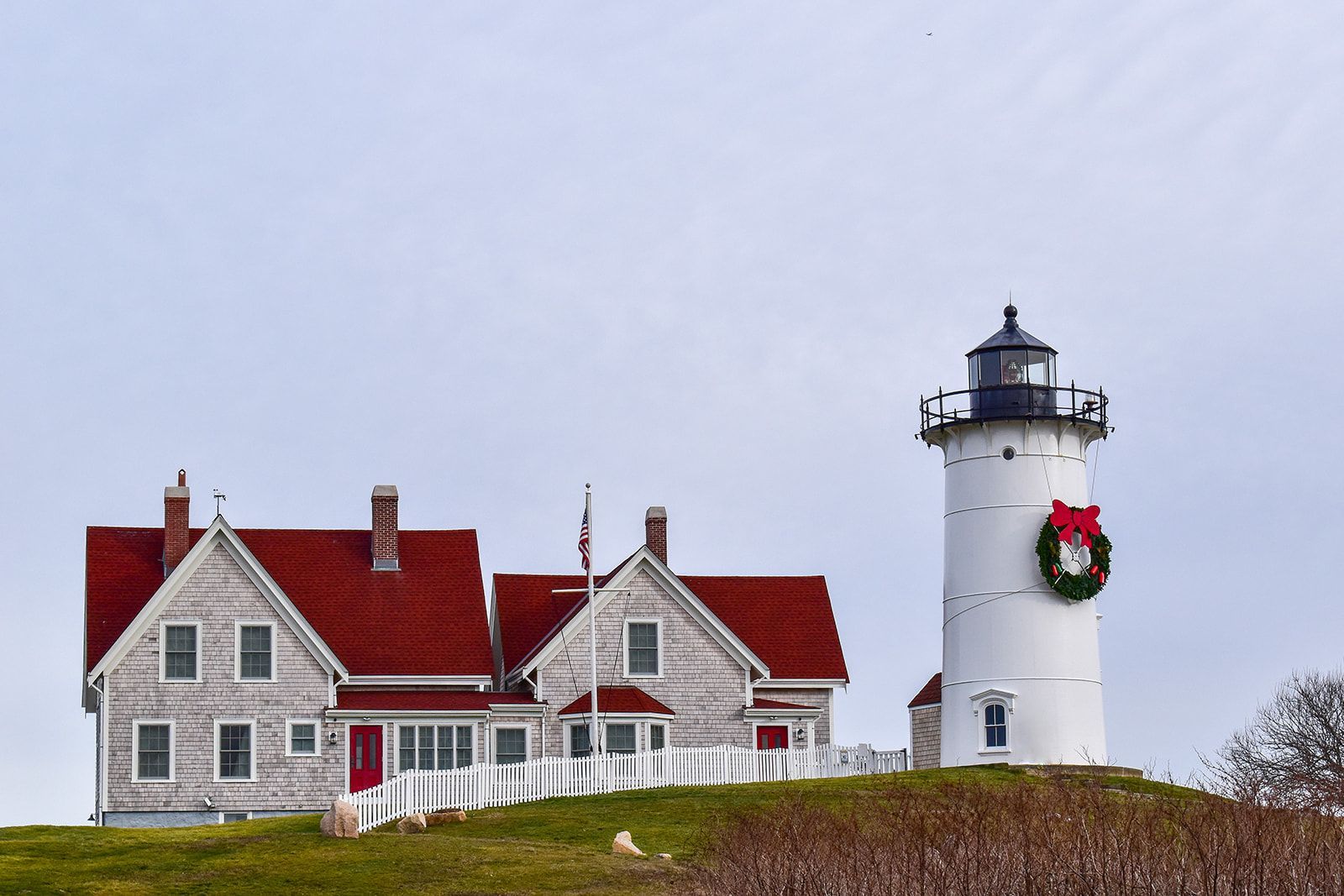 A white lighthouse and an attached house with a red roof on a grassy hill, decorated with a festive holiday wreath.