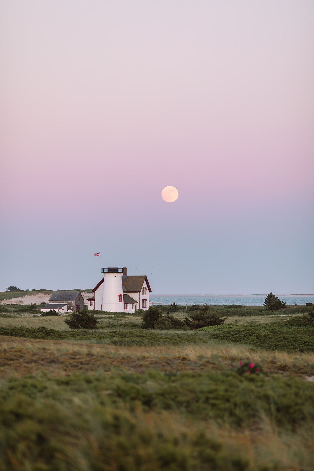 A white lighthouse stands on a grassy coastal landscape under a pastel pink and blue sky with a full moon.