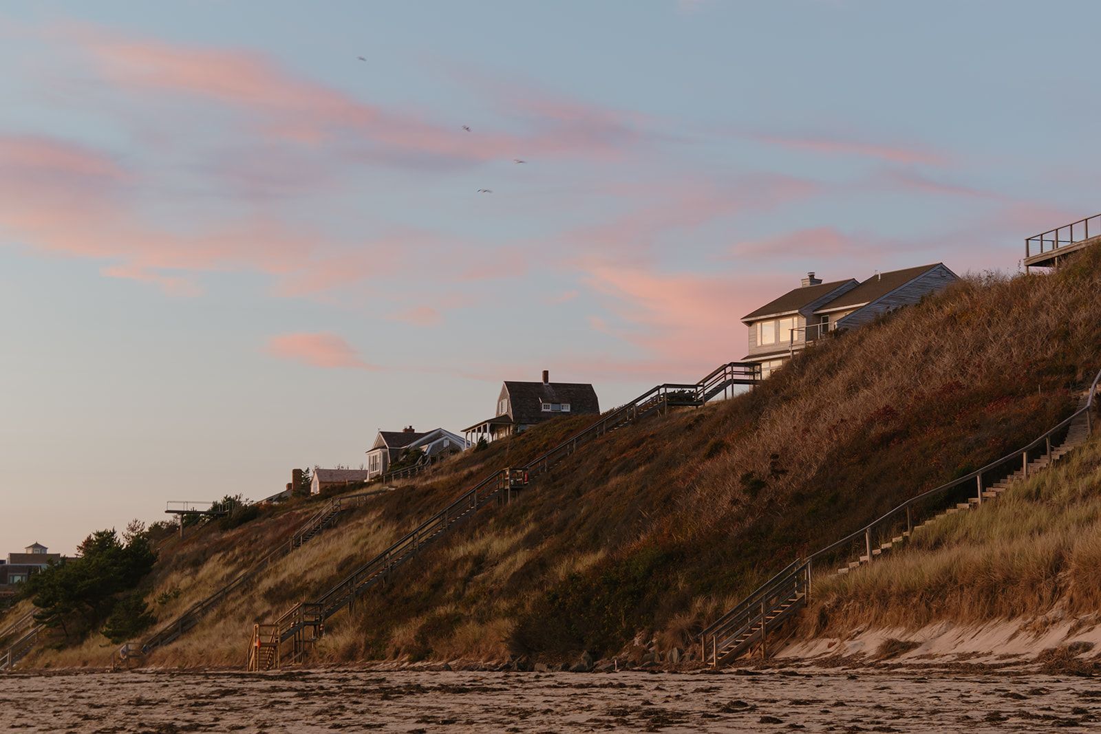 Houses sit atop a grassy bluff overlooking a beach at sunset, with a wooden staircase leading down to the sand.