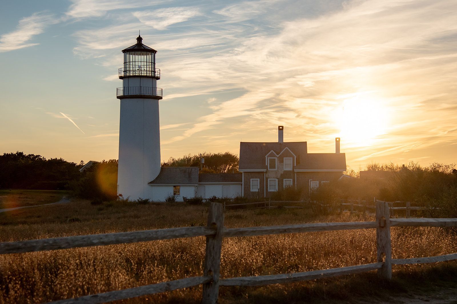 A white lighthouse stands beside a house in a grassy field at sunset, framed by a rustic wooden fence in the foreground.