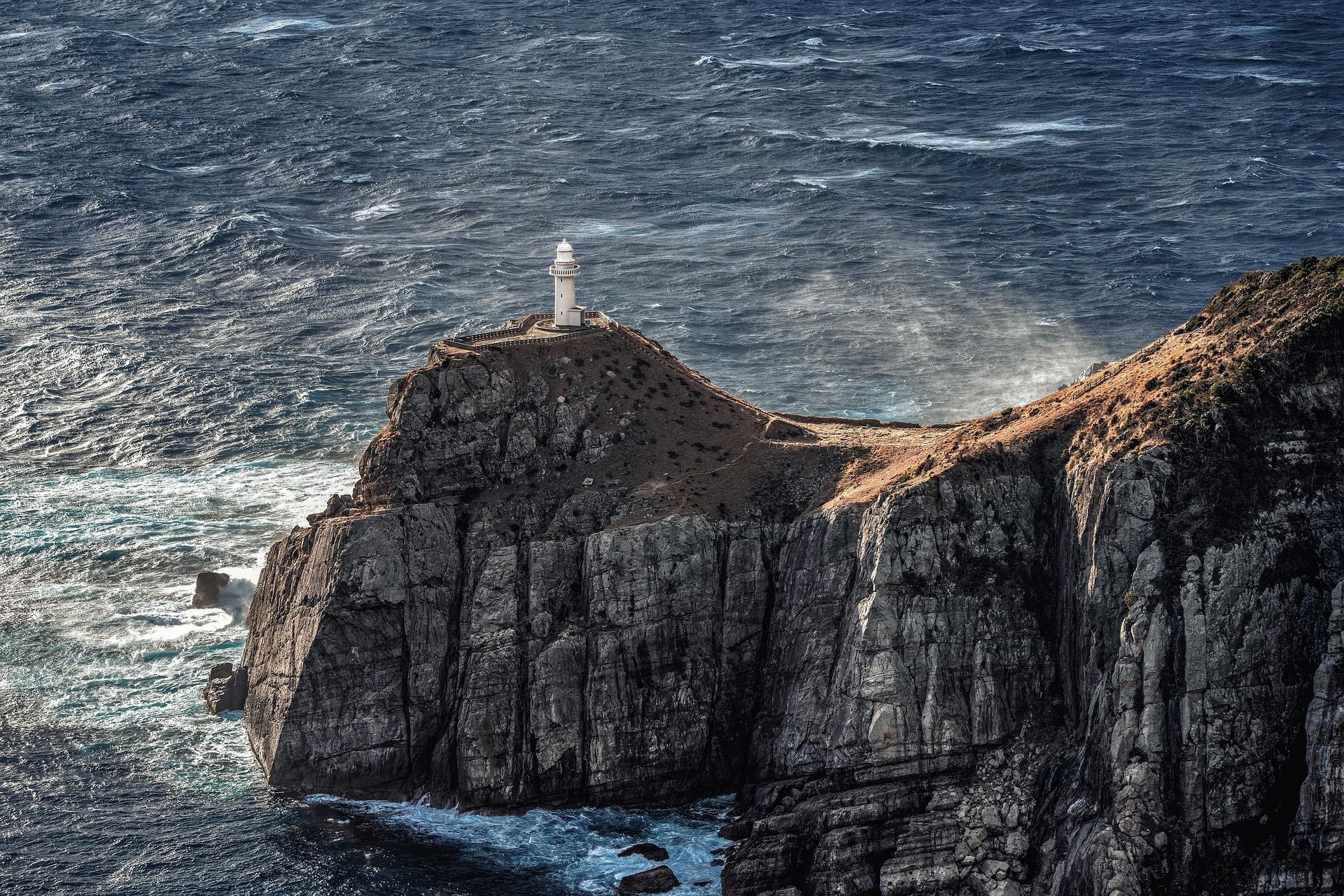 A lighthouse stands on a jagged, dark cliff overlooking a deep blue, churning sea.
