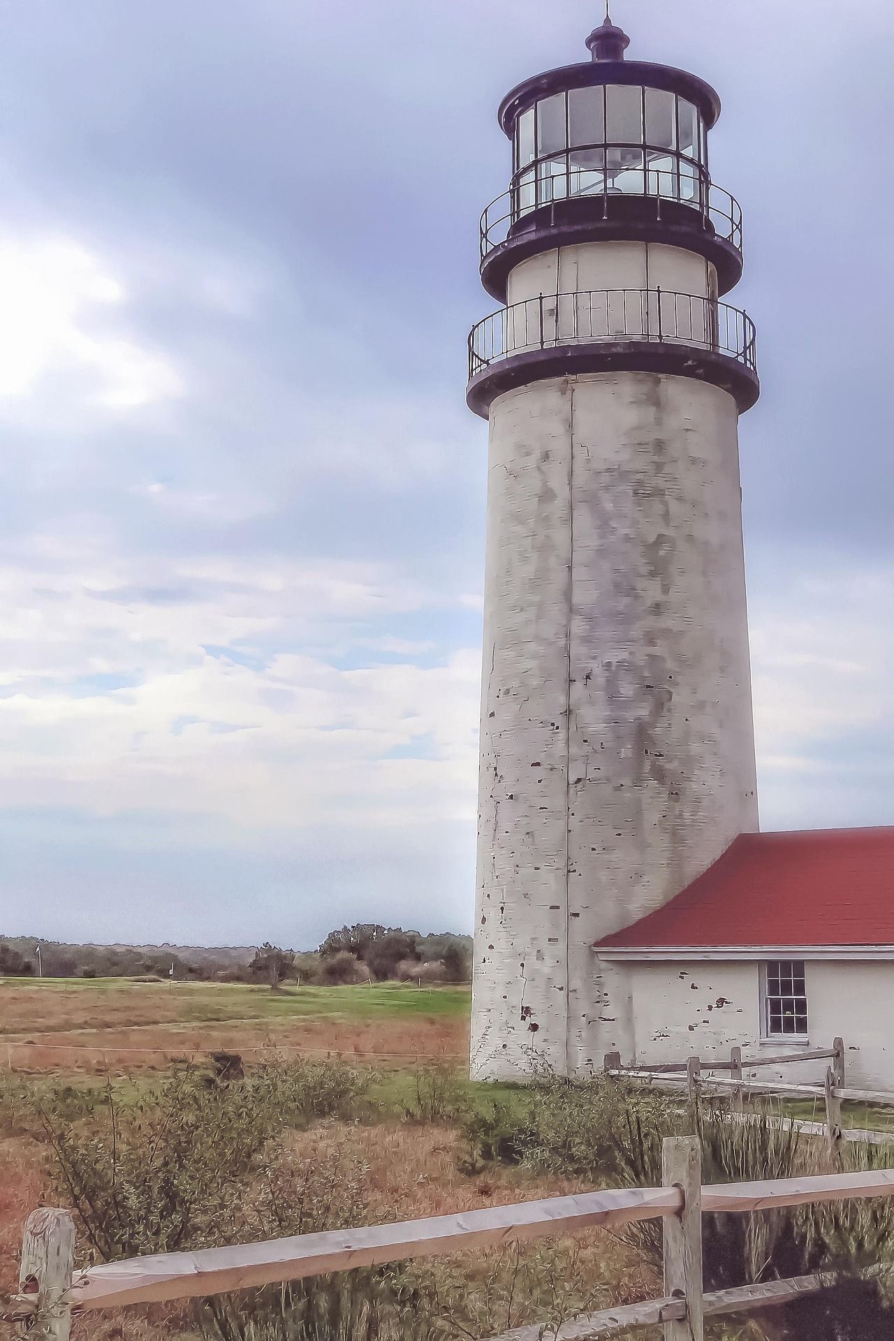 A white, weathered lighthouse stands against a cloudy sky beside a small building with a red roof, fronted by a fence.