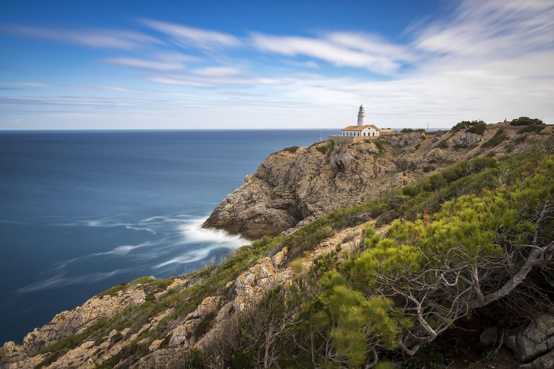 A lighthouse sits atop a rugged rocky cliff overlooking the deep blue sea under a sky with wispy clouds.