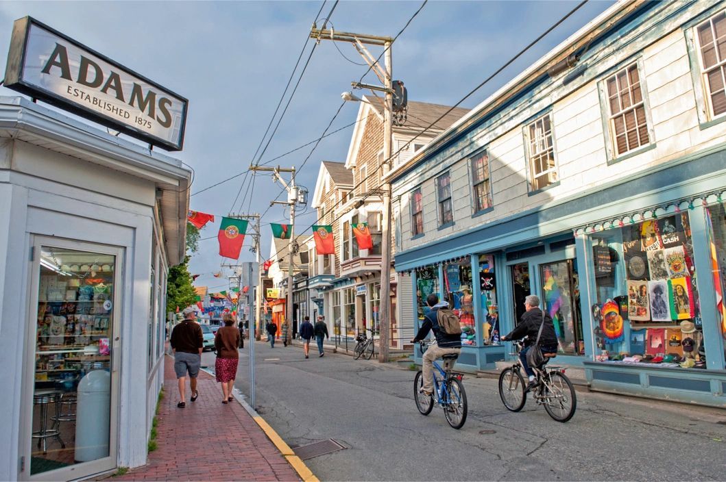 Pedestrians and cyclists travel along a sunlit town street lined with historic shops and hanging flags.