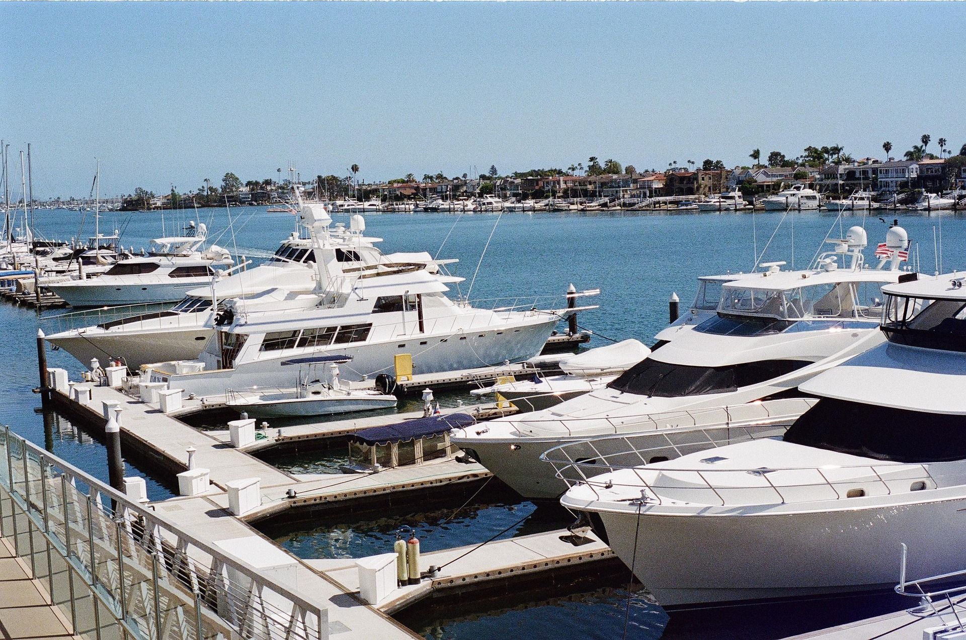 Several white yachts are docked in a sunny harbor lined with waterfront homes.
