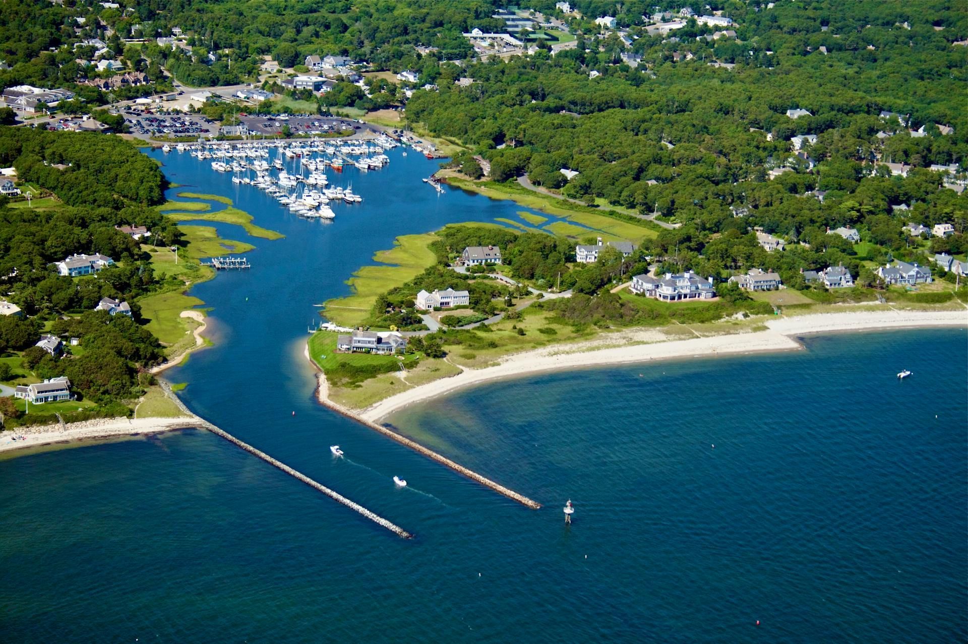 Aerial view of a coastal inlet with stone jetties, a small boat marina, and surrounding houses nestled in green trees.