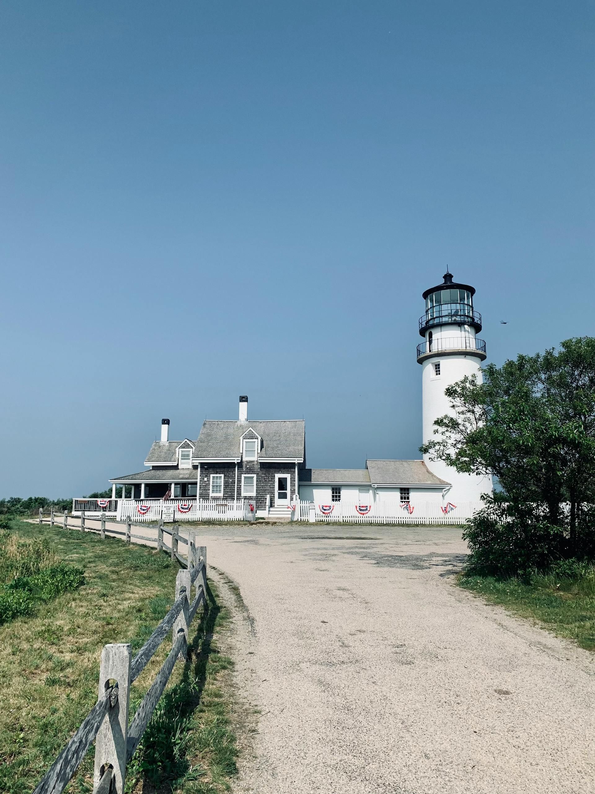 A white lighthouse tower connected to a grey shingled house, set beside a dirt path with a wooden fence under a clear sky.