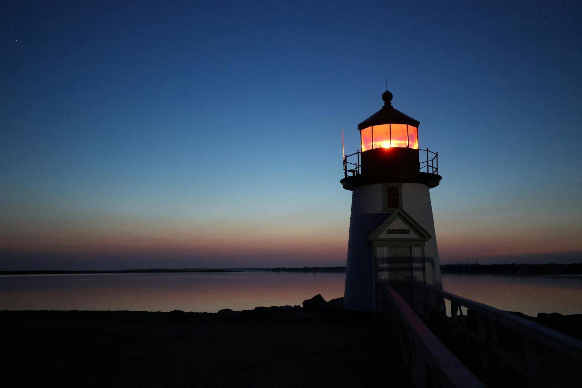 A glowing lighthouse stands on a pier overlooking a calm body of water at twilight, with a gradient sunset sky behind it.