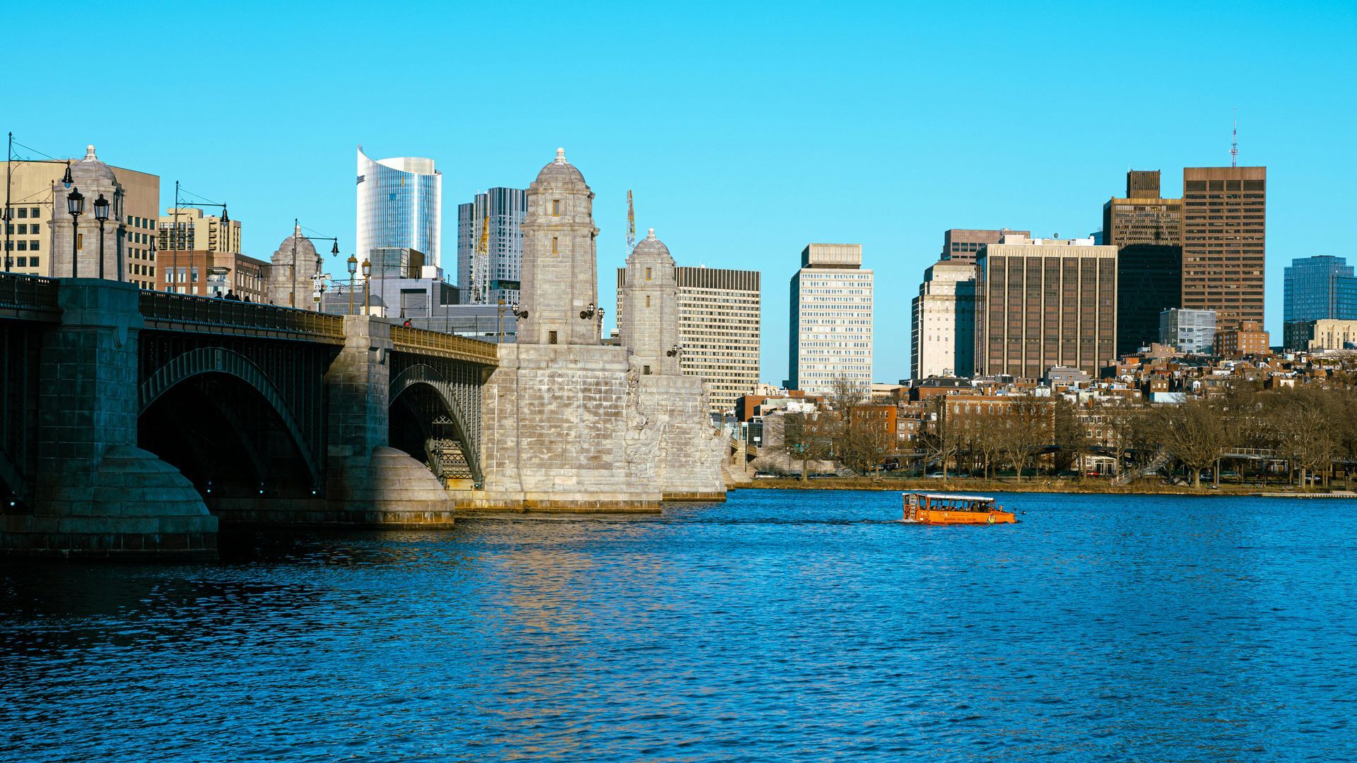 The Longfellow Bridge crosses the Charles River, showing Boston's skyline with a small boat in the foreground.