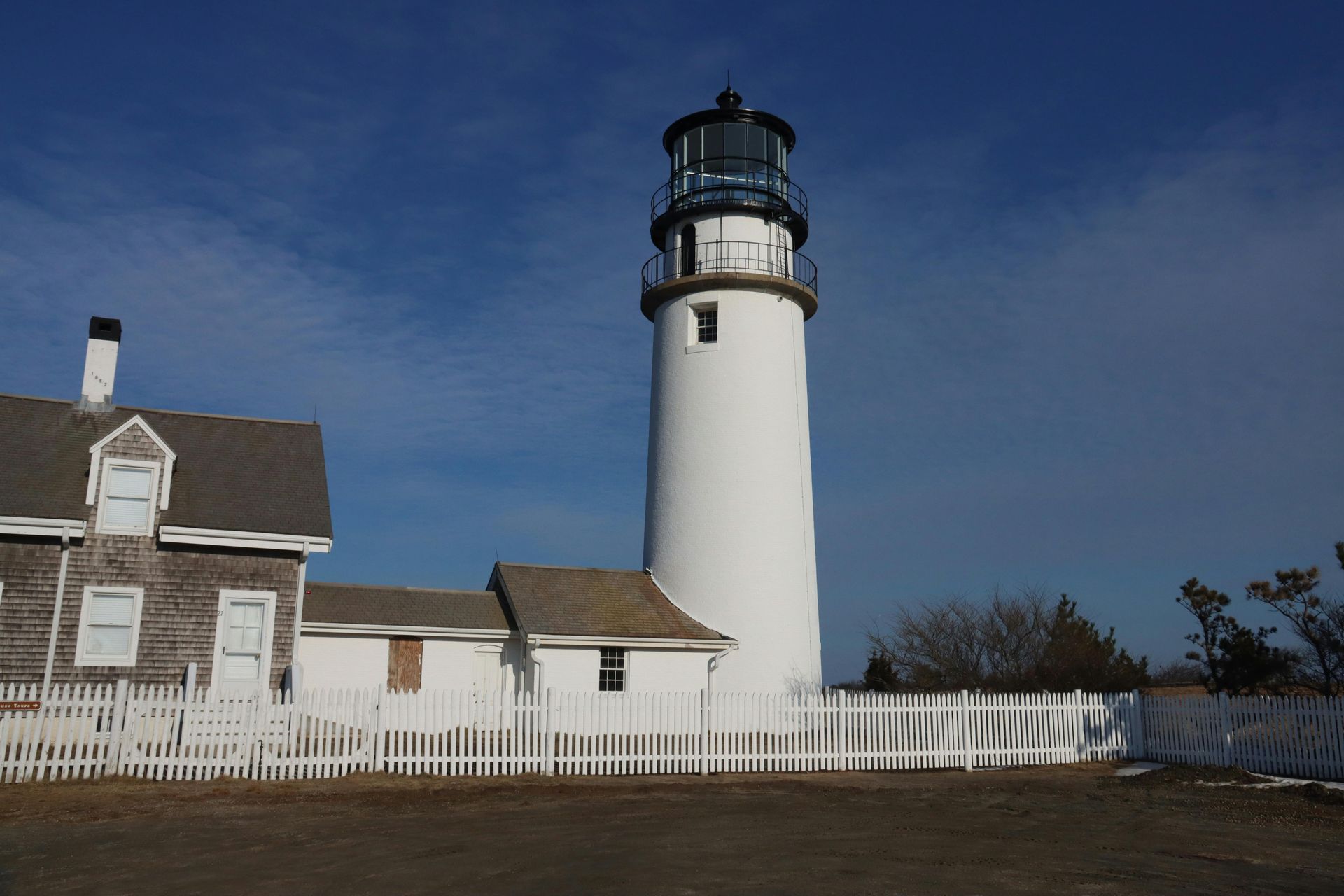 A white lighthouse tower stands beside a shingled building behind a white picket fence under a clear blue sky.