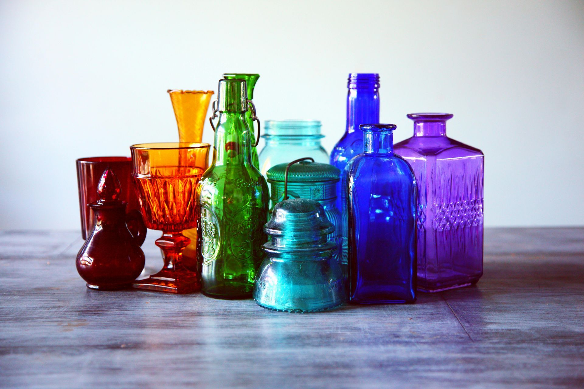 A colorful collection of vintage glass bottles and jars in red, orange, green, blue, and purple on a wooden surface.