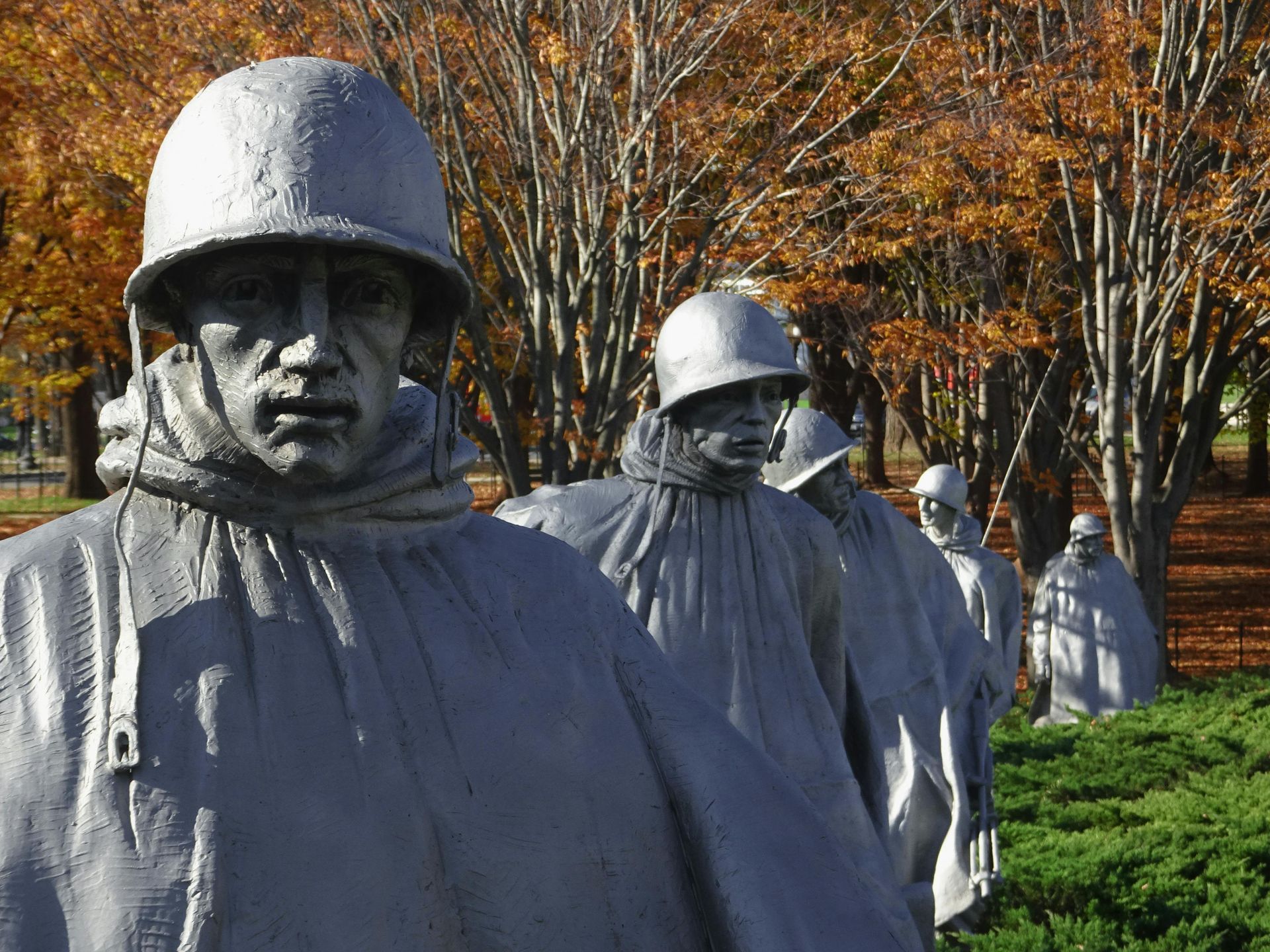 Silver soldier statues in ponchos and helmets stand in a line against an autumn background at the Korean War Memorial.