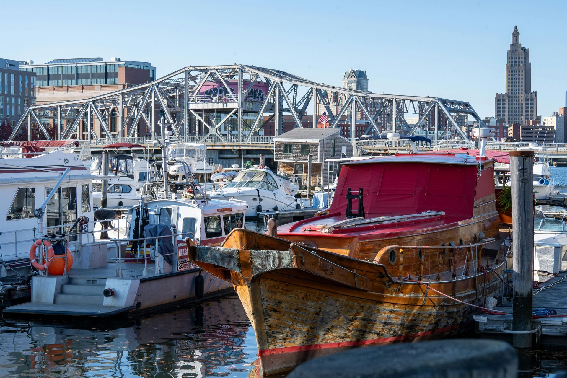 A wooden boat with a red cover in a harbor, with a metal bridge and the Providence skyline in the background.