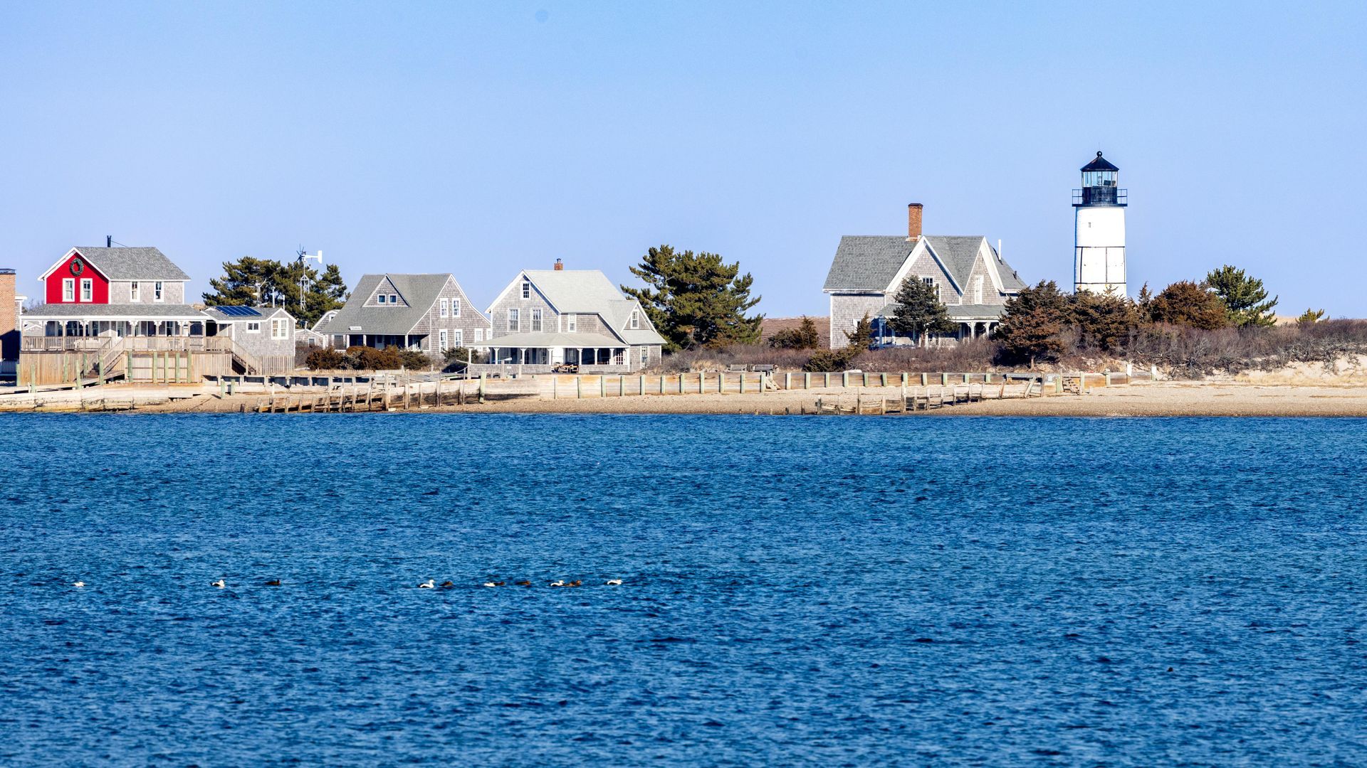 A lighthouse stands on a coastline alongside several houses with shingled siding, overlooking calm blue water.