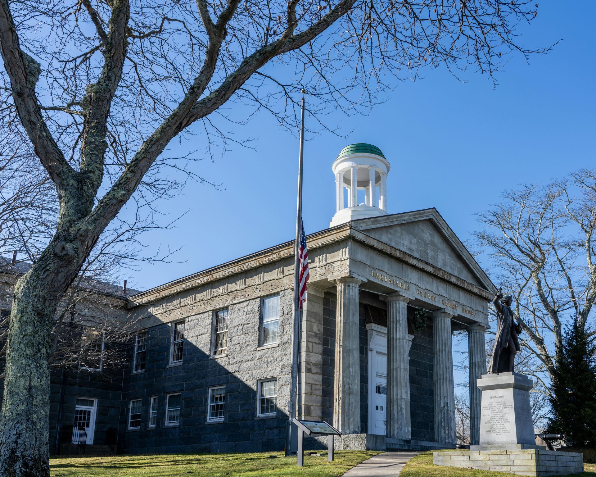 A historic stone building with a white cupola and a front portico, flanked by a flagpole and a statue on a stone base.