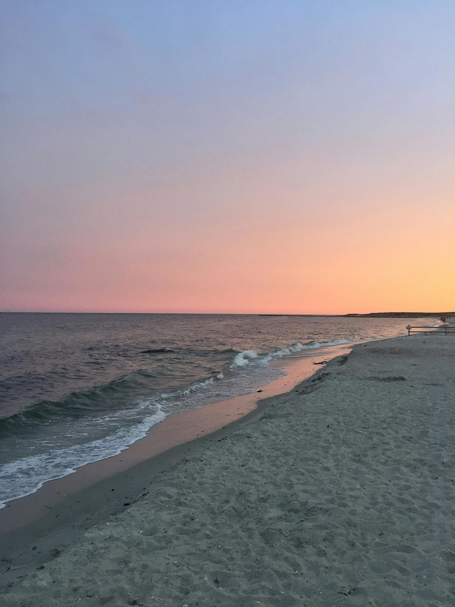 A calm beach at sunset with soft pink and orange hues in the sky reflecting on the gentle waves and wet sand.
